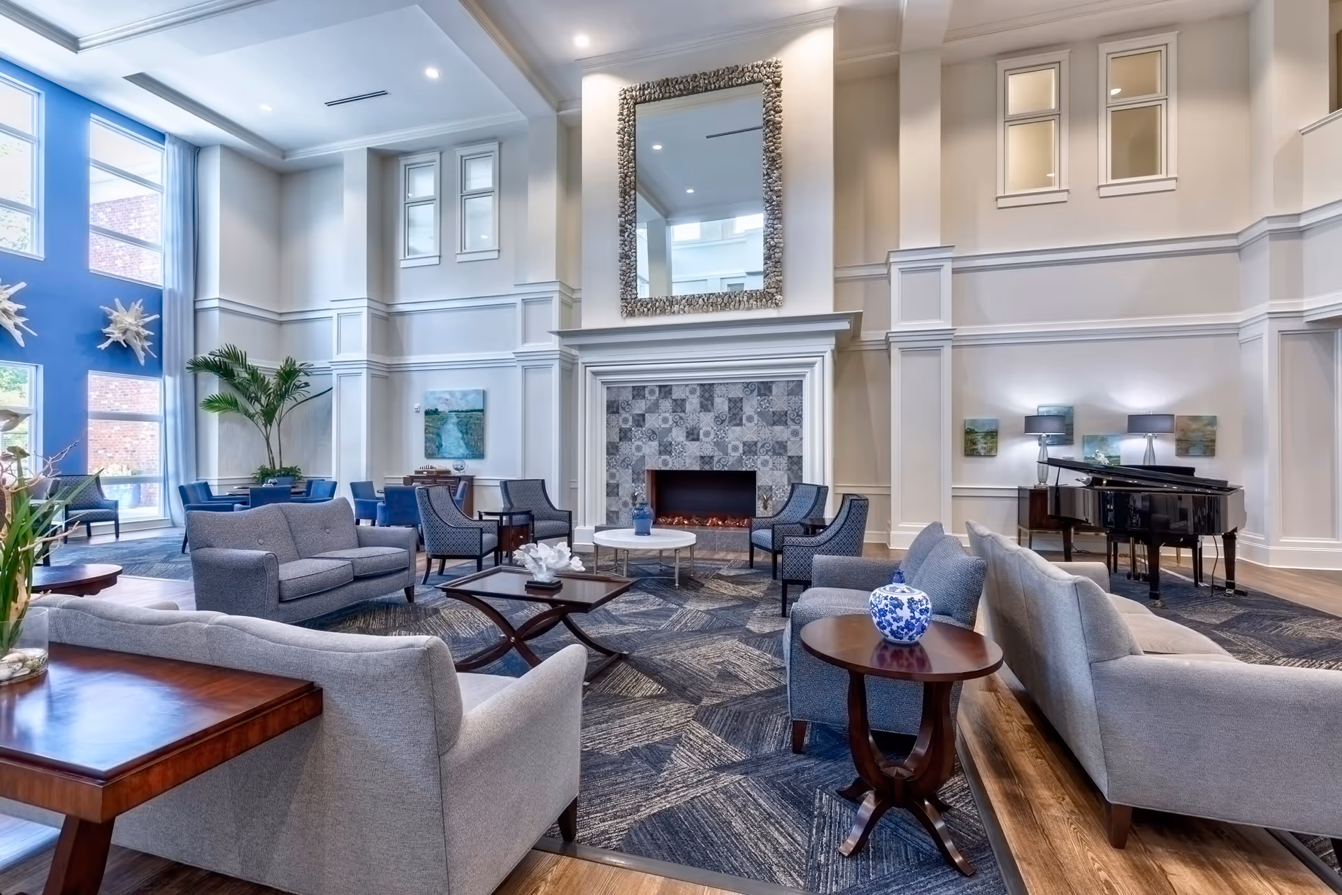 A spacious and elegant living room area in Indigo Hall Assisted Living and Memory Care featuring multiple gray sofas and armchairs arranged around a central fireplace with a decorative tile surround and a large mirror above it. The room has high ceilings, large windows letting in natural light, a grand piano in the corner, blue accent walls, and various tables with decorative items and plants.