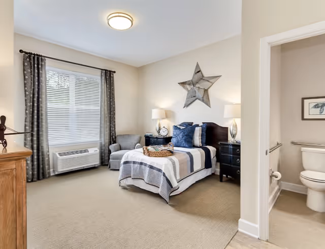 Well-lit bedroom with a single bed dressed in striped bedding, two nightstands with lamps, an armchair by a window, and an open door leading to an ensuite bathroom.