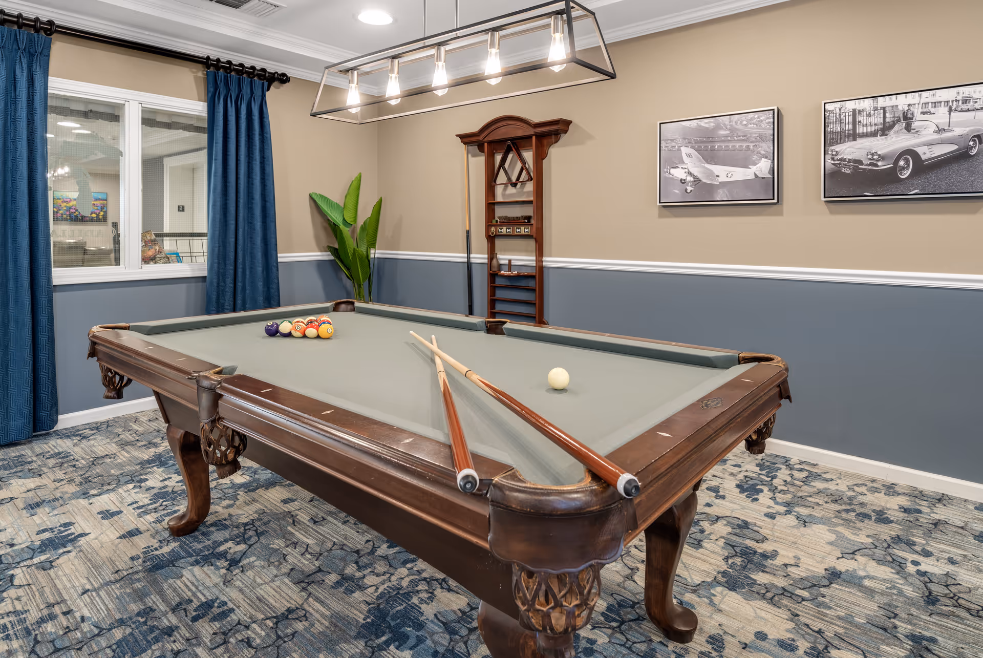 A well-lit game room featuring a classic wooden pool table with two pool cues resting on its edge. The pool table is set with a triangle rack of billiard balls and a white cue ball. The room has blue and beige walls with a white chair rail, blue curtains covering a window, a plant in the corner, and two black-and-white framed photos of vintage vehicles on the wall. The floor is covered with a patterned carpet in shades of blue and gray.