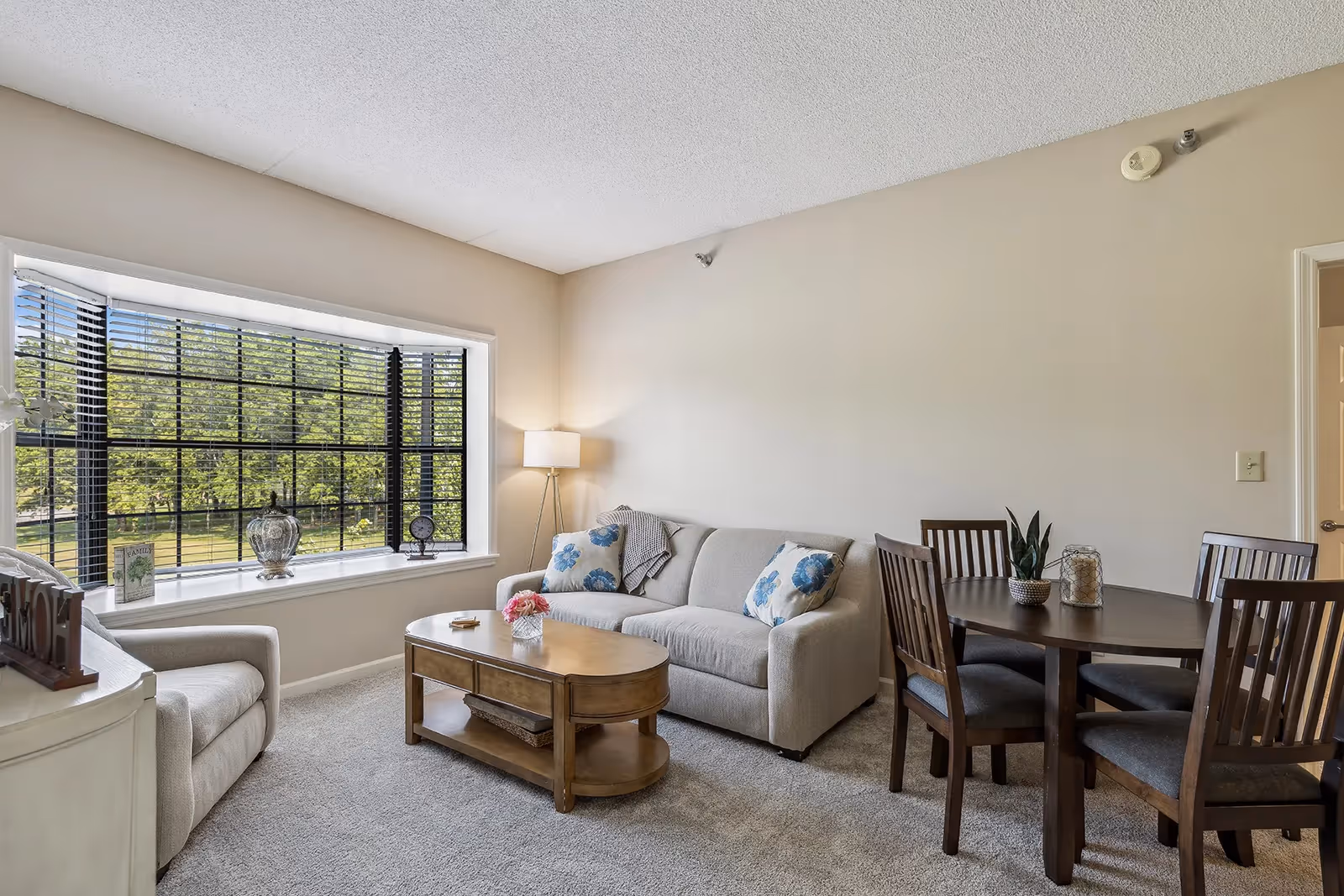 A bright living room with a large window featuring black blinds overlooking greenery outside. The room has a beige sofa with blue floral pillows, a wooden coffee table with a small flower arrangement, a beige armchair, a floor lamp, and a round wooden dining table with four chairs. The walls are painted light beige and the floor is carpeted.
