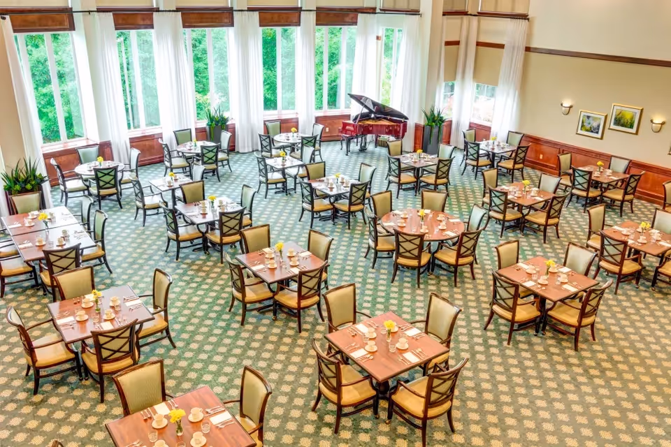 Spacious senior living dining room with multiple set tables, large windows, and a grand piano.