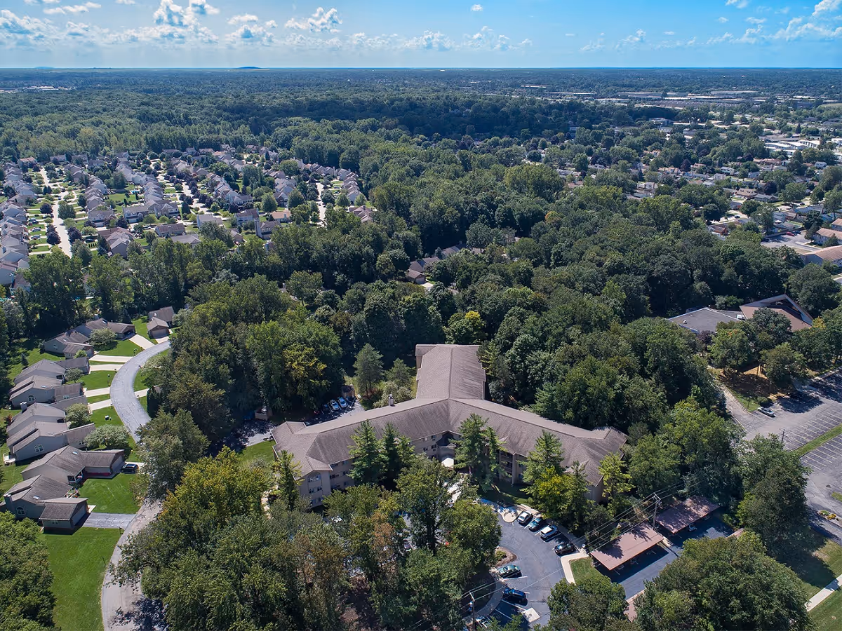 Aerial view of a senior living facility named American House Westland Joy surrounded by dense trees and greenery. The building has a Y-shaped layout with a parking lot and cars visible. Residential houses and neighborhoods are seen in the background under a partly cloudy sky.