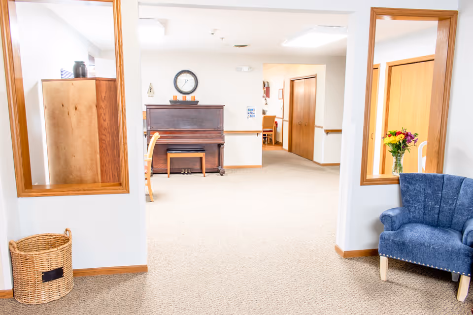Bright senior living common area featuring a piano against the far wall, a blue armchair, a woven basket, and interior pass-through windows.