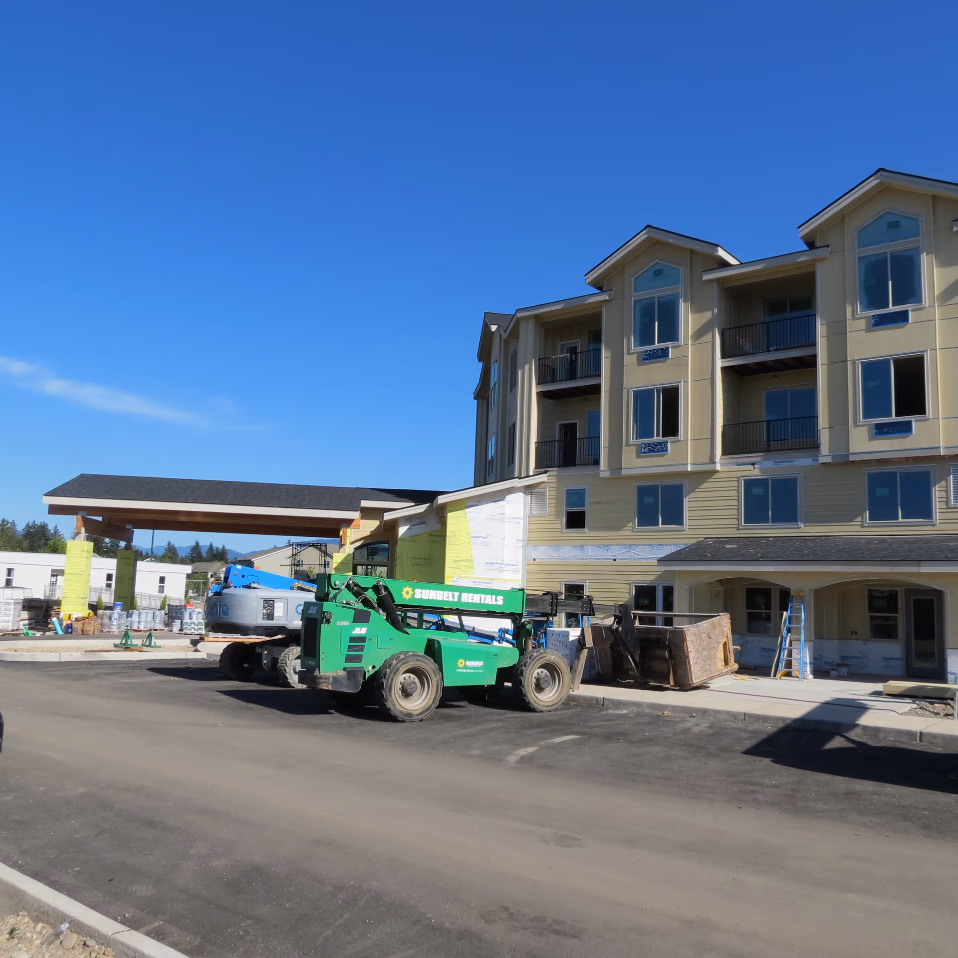 Three-story building front under construction with construction equipment and vehicles parked in front under a clear blue sky.