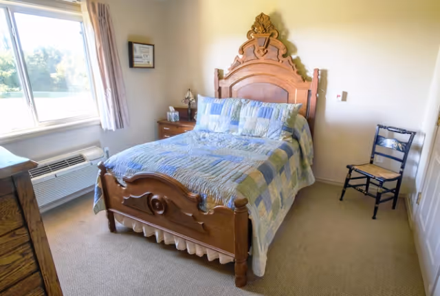 A bedroom with a wooden bed frame featuring an ornate headboard, a patchwork quilt on the bed, a wooden nightstand with a lamp and tissue box, a window with curtains, a wooden dresser, and a simple wooden chair against the wall.