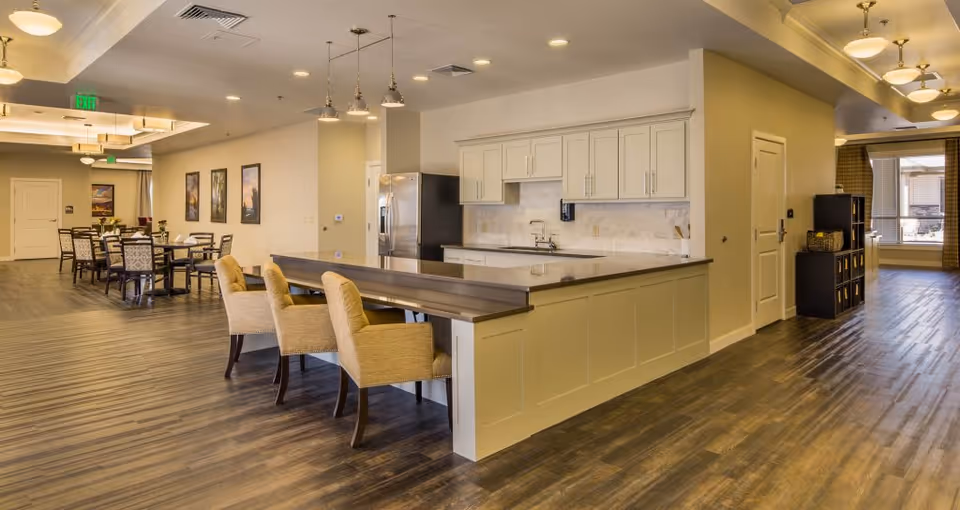 A spacious, well-lit interior area featuring a modern kitchen with white cabinets, a large countertop, and three beige upholstered chairs at a breakfast bar. In the background, there is a dining area with multiple tables and chairs, framed artwork on the walls, and large windows letting in natural light. The flooring is a dark wood finish, and the ceiling has recessed lighting and hanging pendant lights.