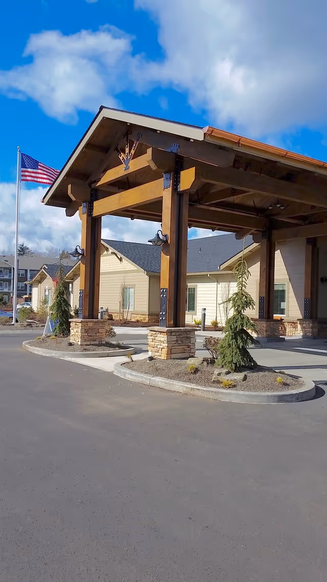Entrance area of Sunnyside Meadows Memory Care facility featuring a large wooden canopy supported by stone pillars, with a paved driveway and small landscaped areas with young evergreen trees. An American flag is visible on a flagpole to the left, and the building exterior is light-colored with a dark roof under a partly cloudy blue sky.