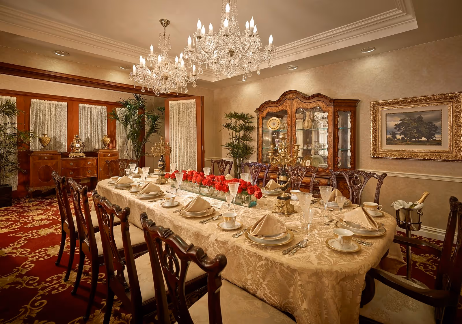 Elegant formal dining room with a long set table, chandeliers, carved wooden chairs, and a china cabinet.