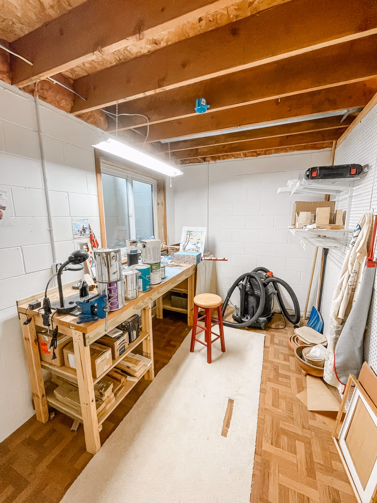 A small workshop or utility room with exposed wooden ceiling beams and white painted cinder block walls. There is a wooden workbench with various paint cans and tools on it, a red stool in the center, a vacuum cleaner in the corner, and shelves with boxes and other items. The floor is partially covered with a light-colored rug, and there is a window on one wall.