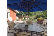Outdoor patio area with a glass-top table and several chairs under a large blue umbrella. The patio is adjacent to a brick building and overlooks a green lawn with trees and shrubs in the background.