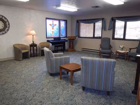 A spacious senior living lounge with striped chairs, a piano, and stained-glass window.