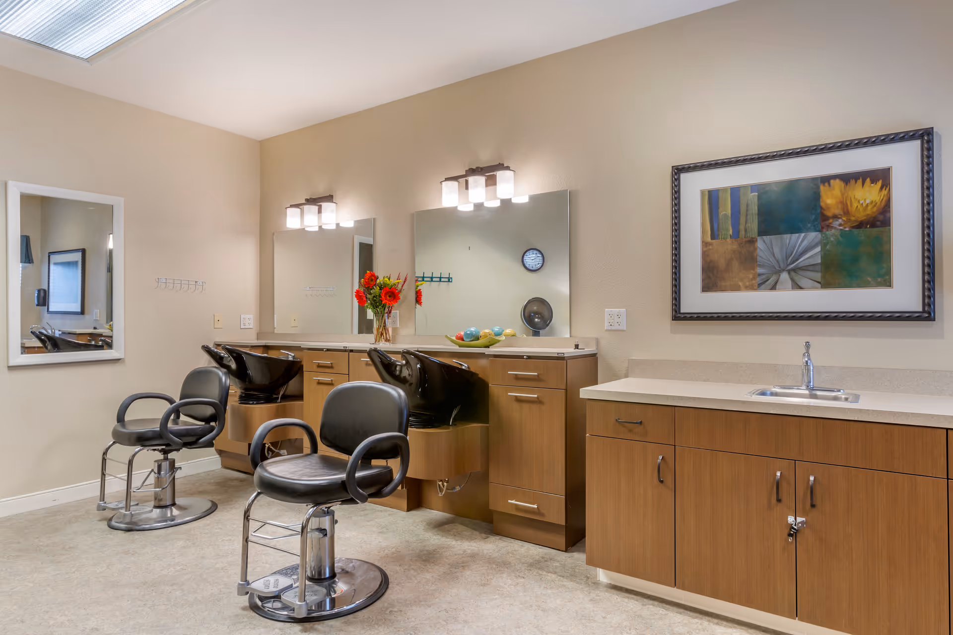 Interior view of a salon area in a senior living facility with two black salon chairs in front of large mirrors mounted on a beige wall. There are wooden cabinets and countertops beneath the mirrors, a vase with red flowers, and a decorative bowl with colorful spheres on the counter. A framed artwork hangs on the wall above a countertop with a small sink.