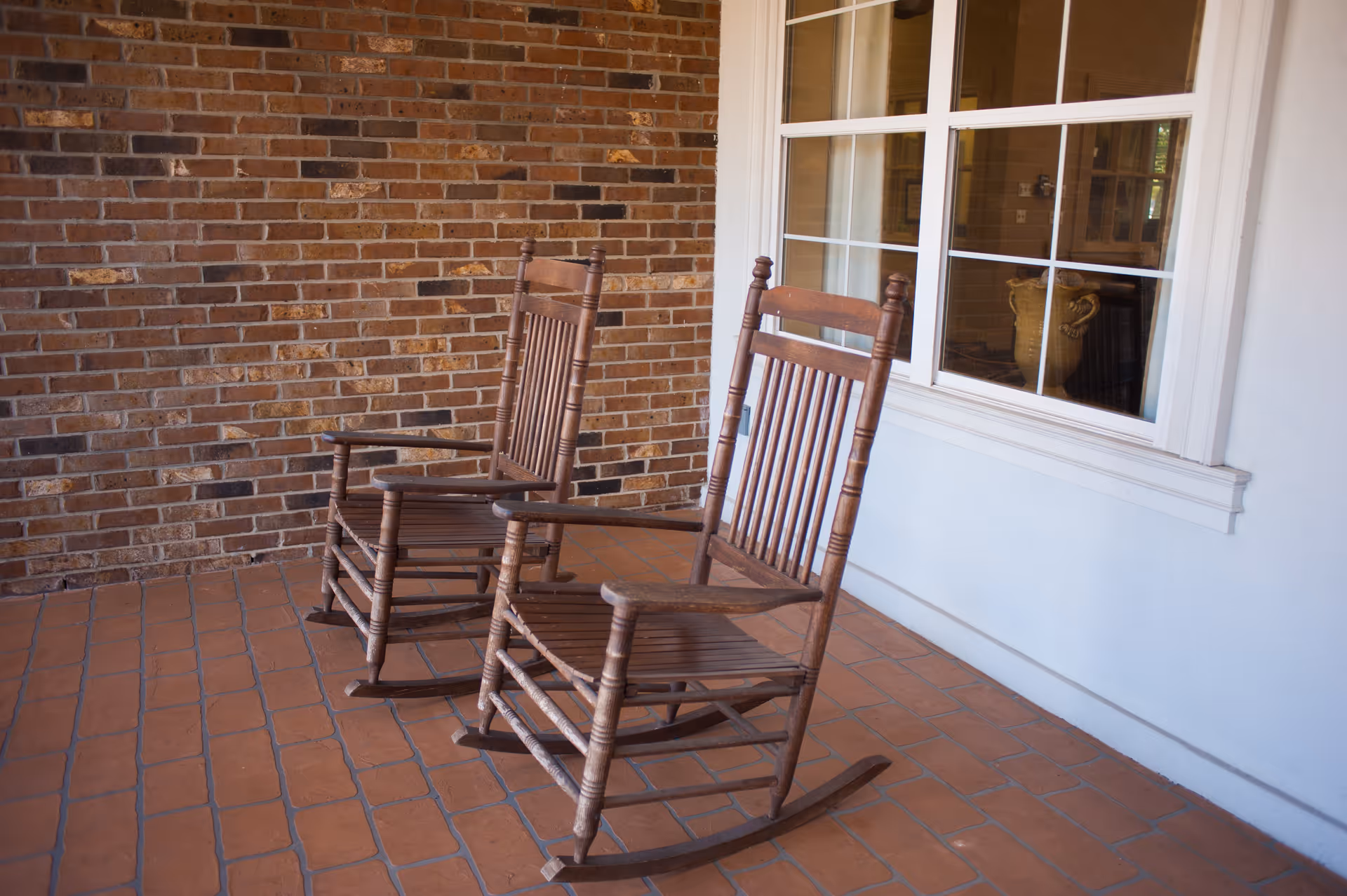 Two wooden rocking chairs on a tiled porch with a brick wall and a window reflecting the interior of a room.