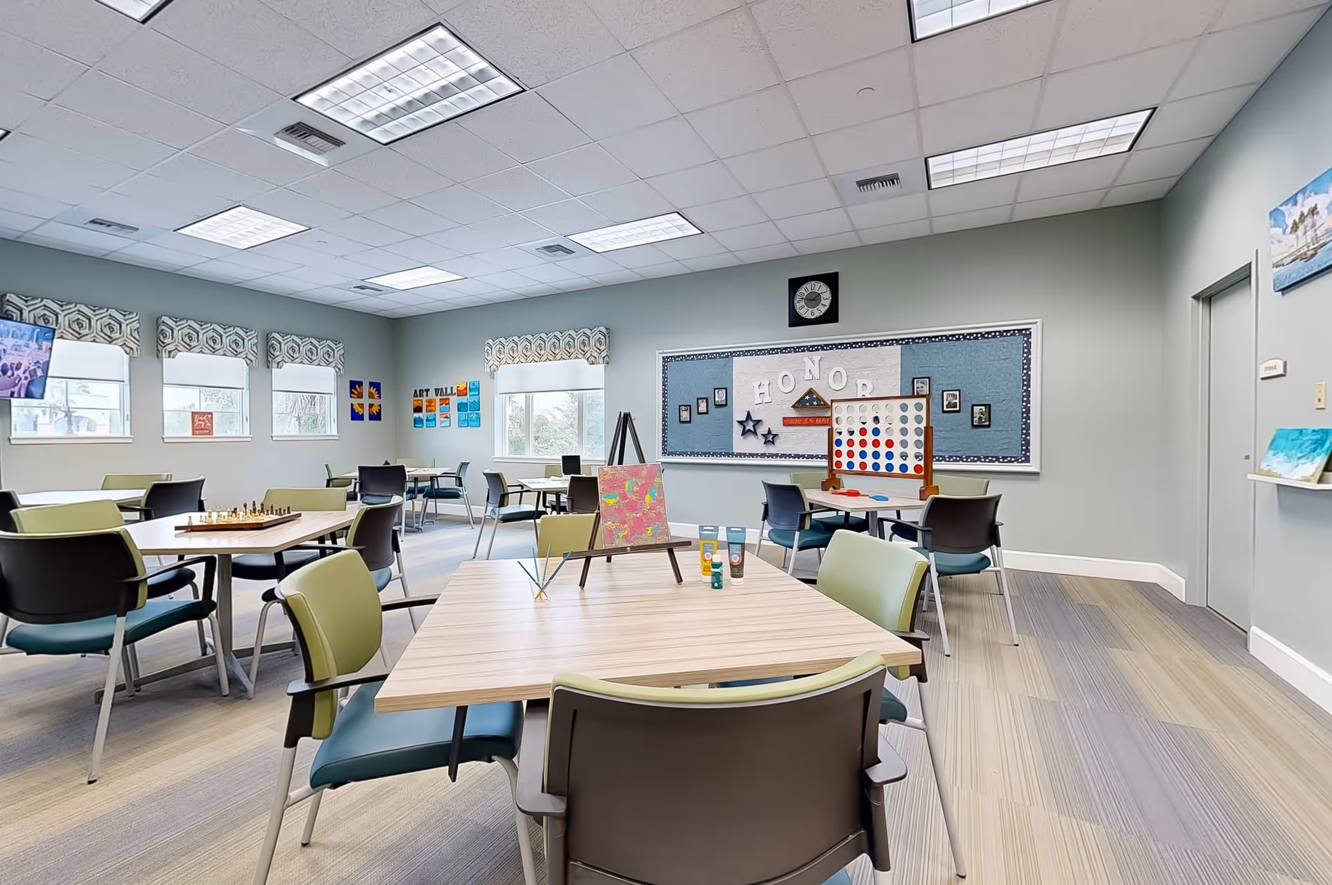 A bright activity room with several tables and chairs arranged for group activities. The room features large windows with patterned valances, a bulletin board labeled 'HONOR' with photos, and various games and art supplies including a chessboard, a large Connect Four game, and painting materials on the tables. The walls are painted light blue and the floor has a light wood pattern.