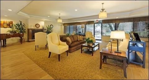 A spacious and well-lit living room area with a brown sofa, two yellow armchairs, a coffee table with a plant, side tables with lamps, and large windows with blinds. The room has a warm color scheme with a patterned carpet and wooden flooring near the entrance. There are decorative plants and wall art in the background.