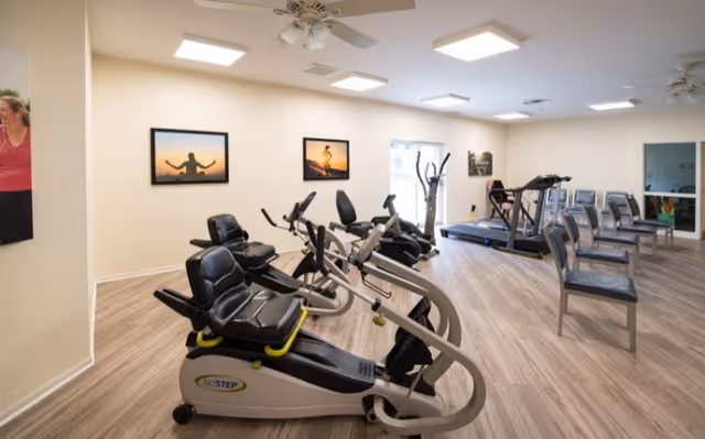 A fitness room with exercise equipment including recumbent bikes, treadmills, and chairs arranged along the wall. The room has light-colored walls, wood flooring, ceiling lights, and framed pictures on the walls.