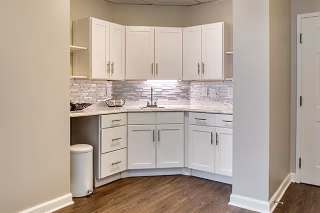 A modern kitchen corner with white cabinets and drawers, a marble countertop, a stainless steel sink, and a tiled backsplash in shades of gray and white. The floor is dark wood, and there is a white trash bin tucked under the counter on the left side.