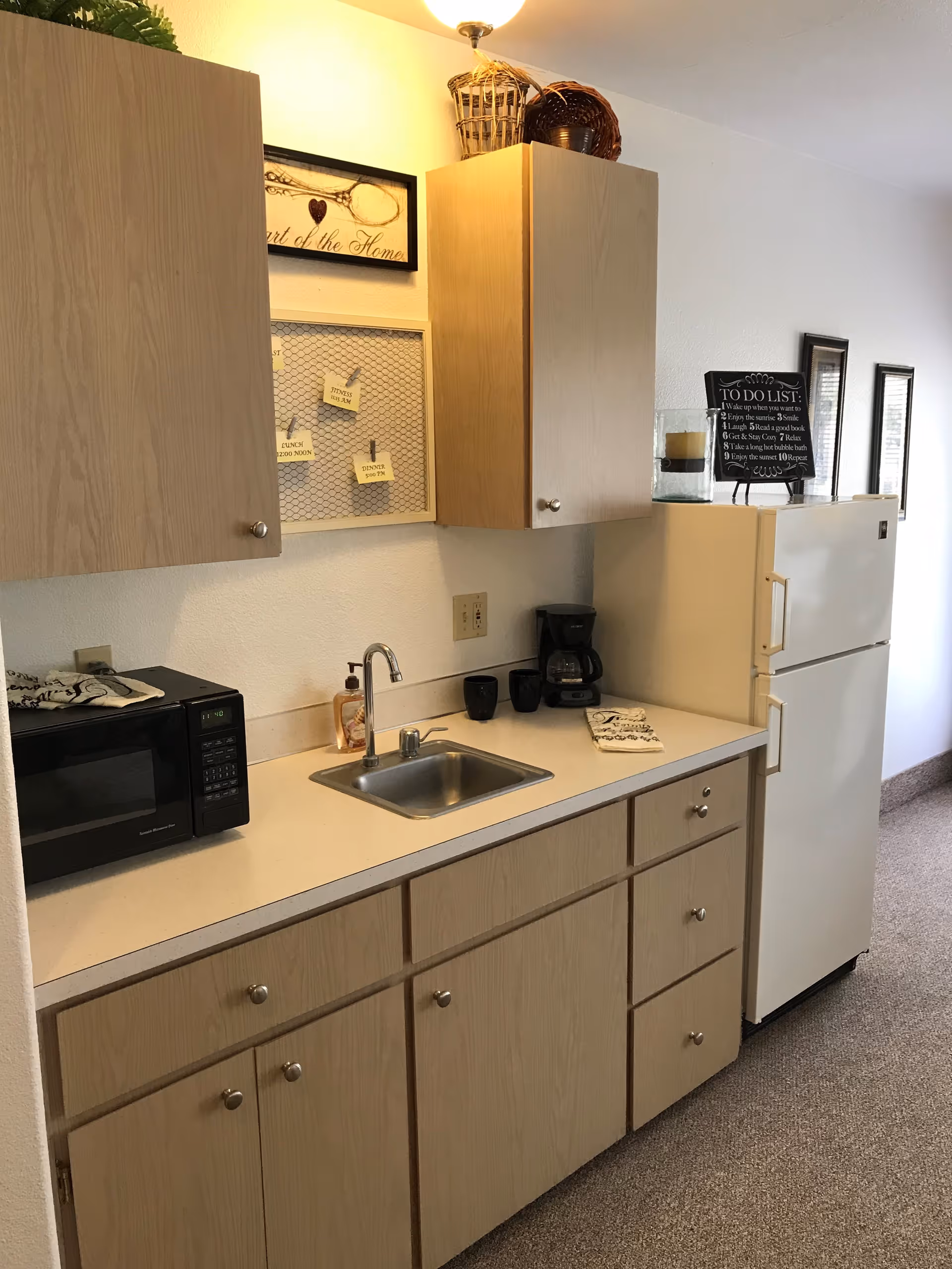 A small kitchen area with light wood cabinets, a white countertop, a stainless steel sink, a black microwave, a coffee maker with two black mugs, and a white refrigerator. Above the cabinets are decorative baskets and framed wall art, including a sign with a to-do list and a framed piece with notes pinned on a wire mesh.