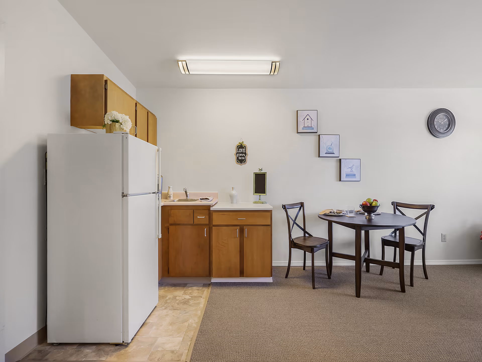 A small kitchen and dining area in an assisted living facility. The kitchen has wooden cabinets, a white refrigerator, a sink, and a countertop with a few decorative items. Adjacent to the kitchen is a round dark wood dining table with two matching chairs. On the wall above the table are three framed pictures arranged diagonally and a round clock. The floor transitions from tile in the kitchen area to carpet in the dining area.