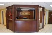 Reception desk area with dark wood paneling and a sign that reads 'Book Walker' mounted on the wall behind the desk. The desk has a computer monitor and a chair behind it. There are hallways visible on both sides of the reception area with beige walls and tiled floors.