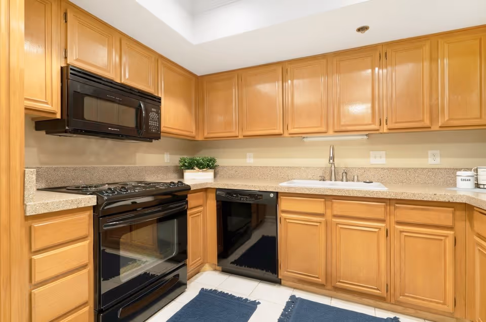 A kitchen with light wood cabinets, black appliances including a stove, microwave, and dishwasher, beige countertops, a white sink with a silver faucet, and two blue rugs on the white tiled floor. There is a small green plant on the countertop and containers labeled 'SUGAR' on the right side.