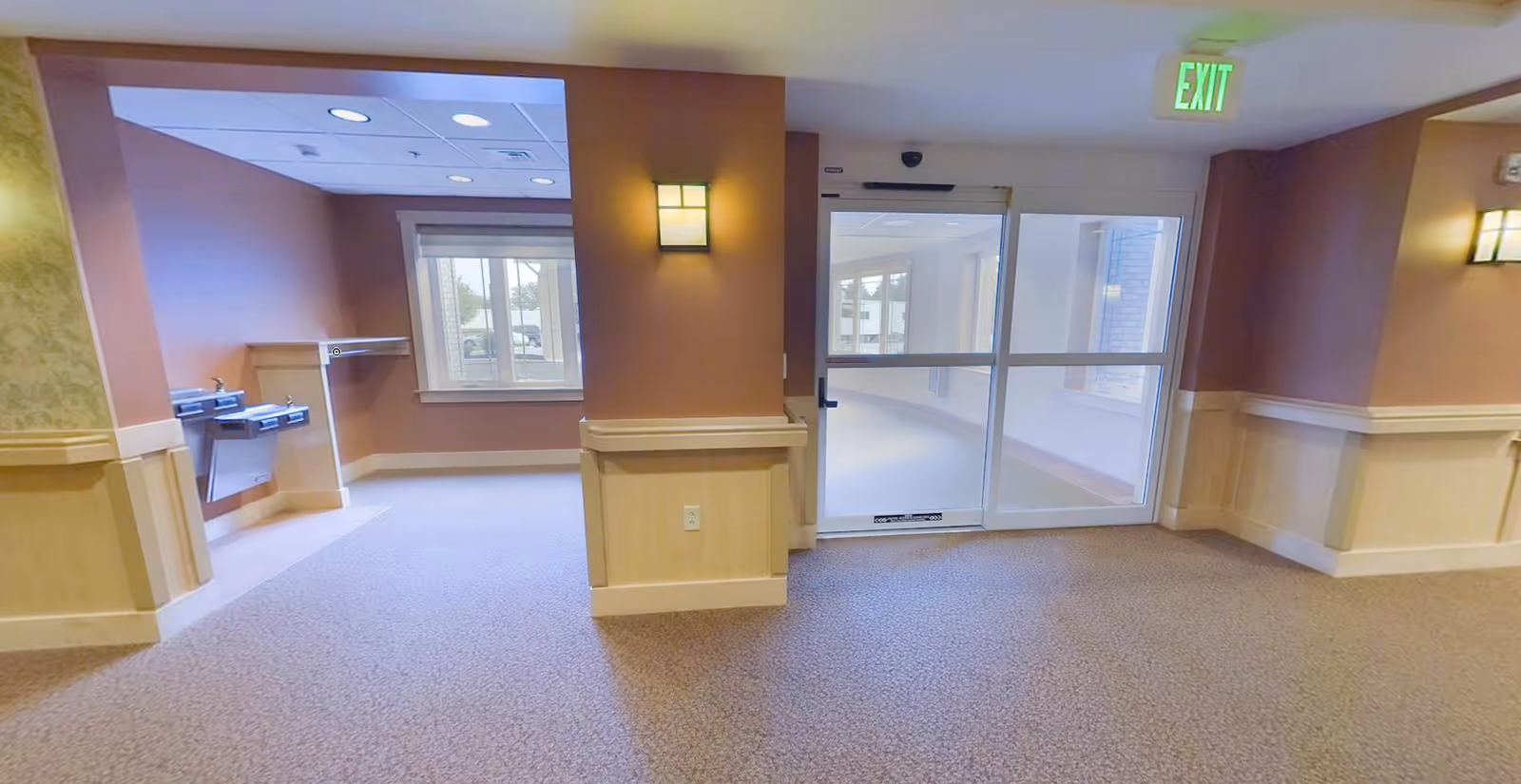 Interior view of a hallway in a senior living facility with beige and brown walls, carpeted floor, two water fountains on the left, a window, a wall-mounted light fixture, and a glass exit door with an illuminated green EXIT sign above it.