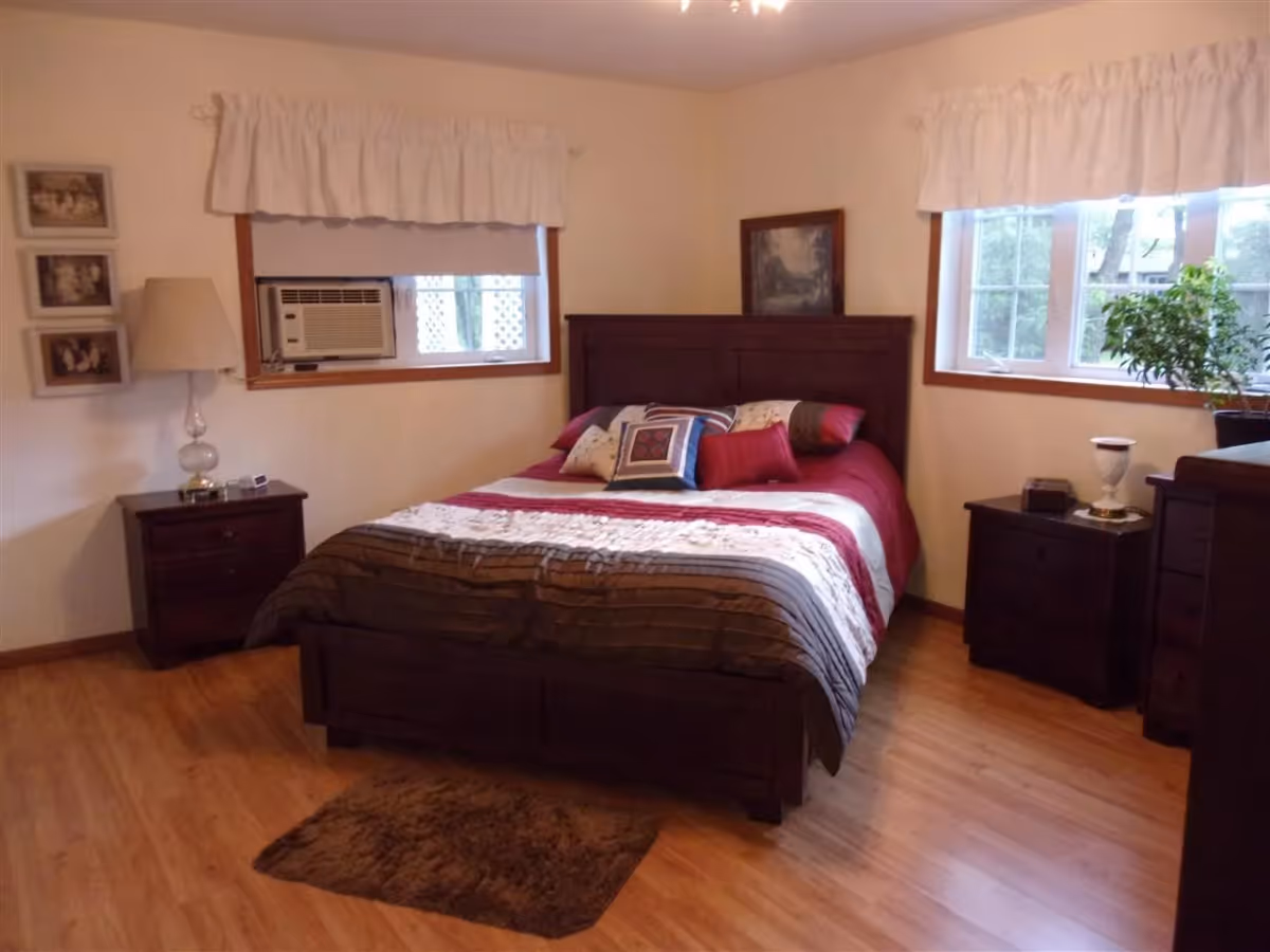 A furnished bedroom with a double bed, two nightstands, windows with valances, and a hardwood floor.