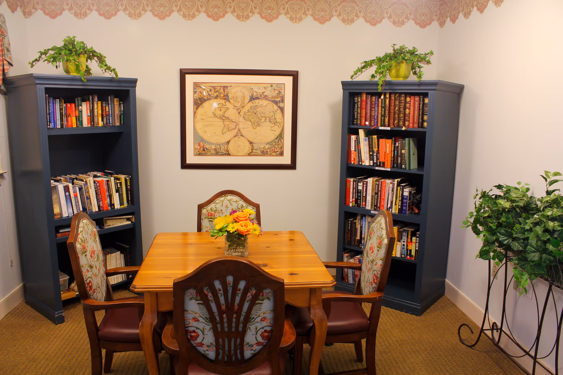 A cozy room with a wooden table surrounded by four upholstered chairs with floral patterns. Two tall blue bookshelves filled with books flank a framed vintage world map on the wall. There are green plants on top of each bookshelf and a larger plant in a decorative stand to the right.