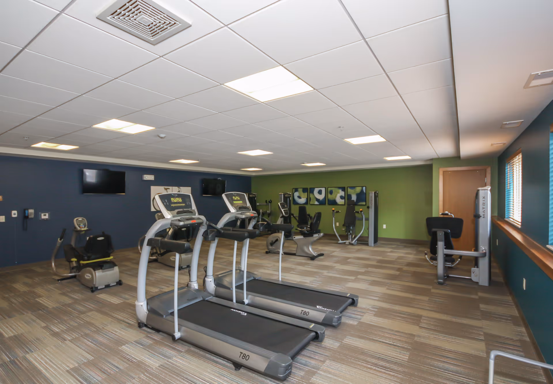 A fitness room with exercise equipment including two treadmills, stationary bikes, and weight machines. The room has a green accent wall with abstract artwork, blue walls, carpeted flooring, and windows with blinds letting in natural light.