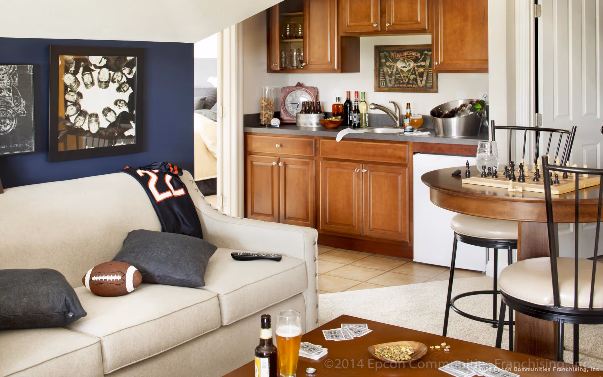 A cozy living area with a beige sofa adorned with dark pillows, a football, and a sports jersey draped over the back. A remote control rests on the sofa. Behind the sofa is a dark blue wall with framed artwork. To the right, there is a small kitchenette with wooden cabinets, a sink, and various bottles and snacks on the counter. In front of the kitchenette is a round wooden table with a chessboard and pieces set up, surrounded by chairs. The floor is tiled near the kitchenette and carpeted in the living area.