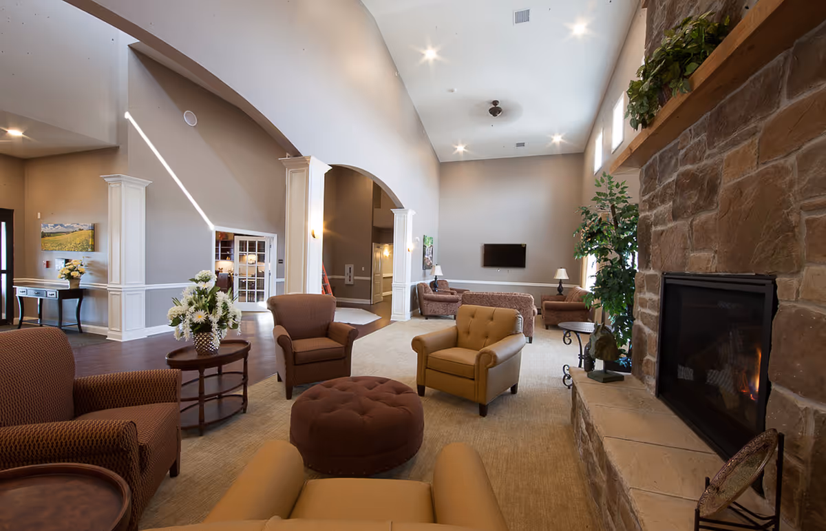 A spacious senior living facility common area with high ceilings, a stone fireplace on the right, several comfortable armchairs and sofas arranged around a central ottoman, a wall-mounted TV, and decorative plants and flowers. The room features neutral tones and soft lighting, creating a warm and inviting atmosphere.