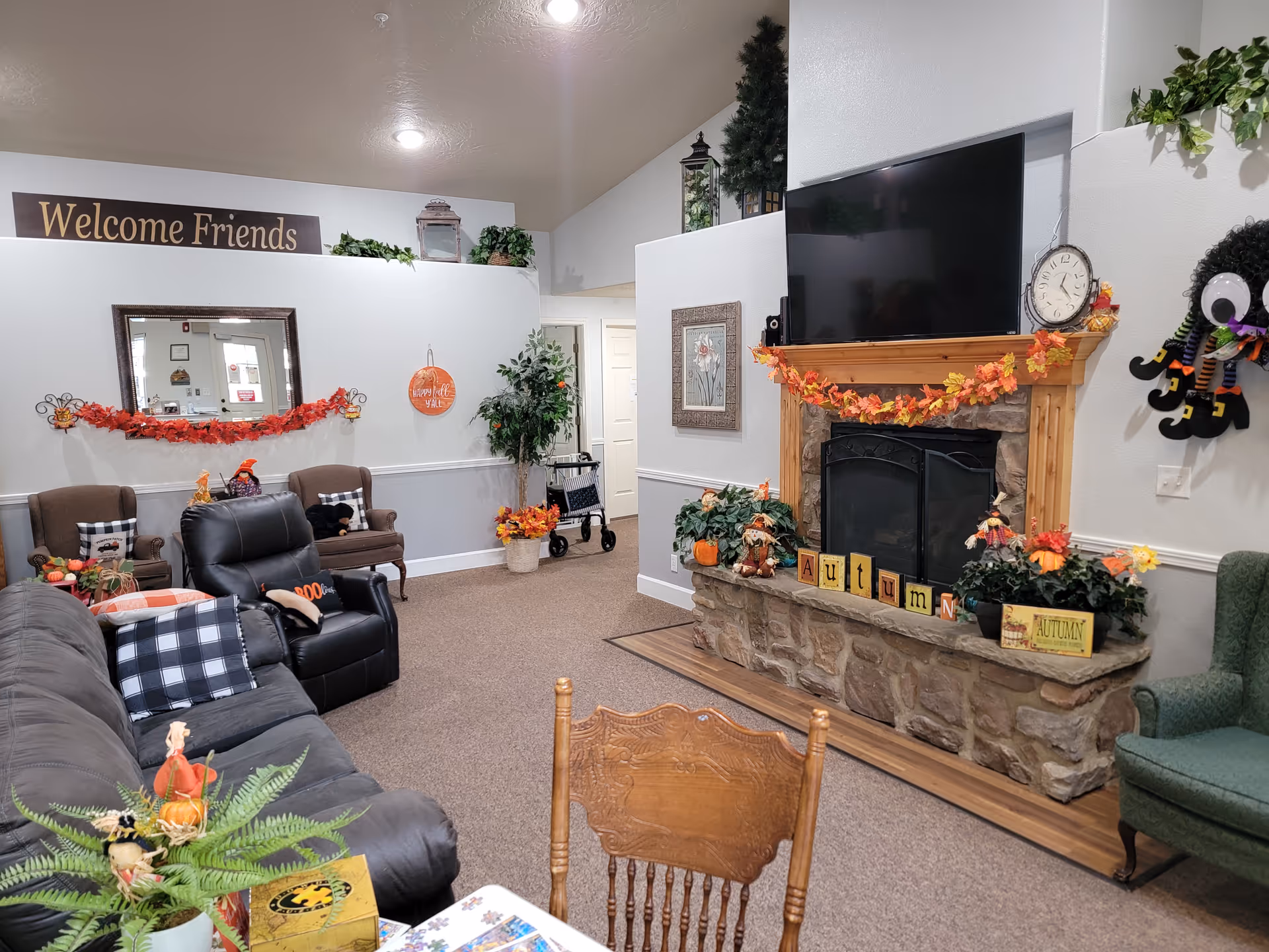 A cozy living room decorated for autumn with a stone fireplace adorned with fall leaves garland and seasonal decorations. Above the fireplace is a large flat-screen TV and a clock. The room features a gray couch with plaid pillows, a black recliner, two brown armchairs, and a green armchair. A wooden chair and table with a plant and puzzle are in the foreground. A sign on the wall reads 'Welcome Friends' and there are various plants and fall-themed decorations throughout the space.