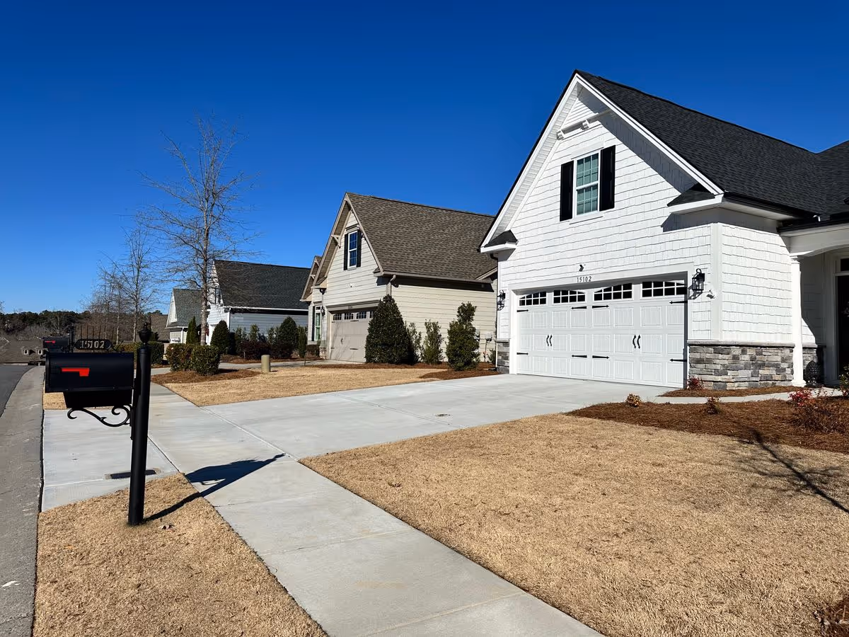 Front exterior of suburban houses showing a driveway, garage, mailbox, and lawn under a clear blue sky.
