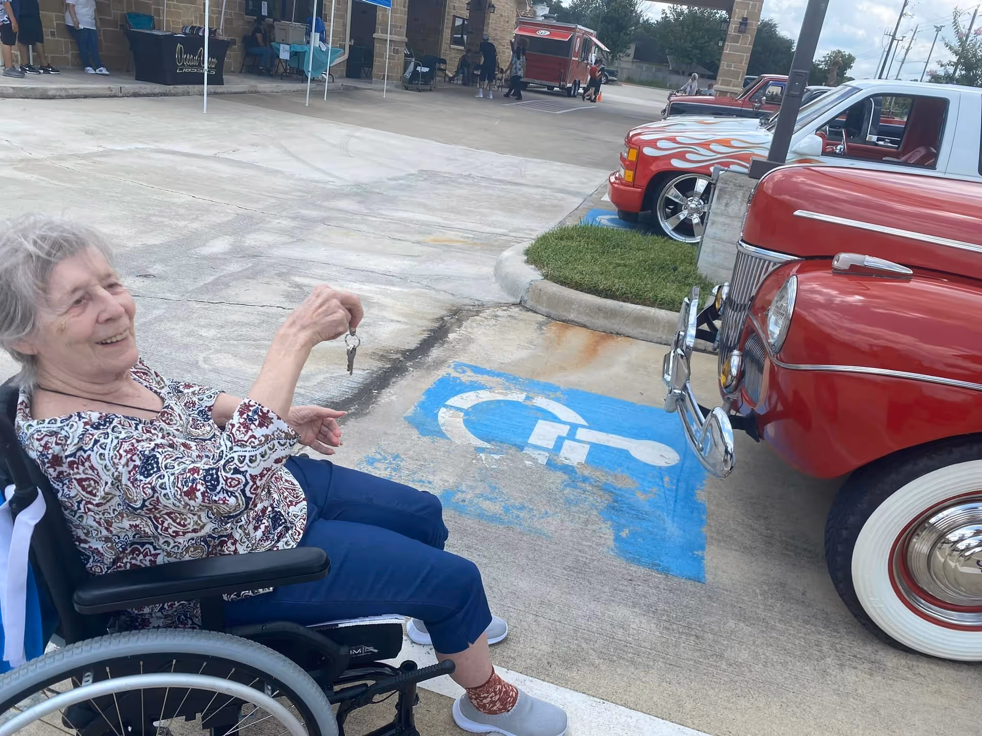 An elderly woman in a wheelchair smiling and holding a set of keys in an outdoor parking area near a blue handicapped parking symbol painted on the ground. In the background, there are vintage cars, including a red car with whitewall tires and a red truck with flame decals, as well as a building with people and tents.