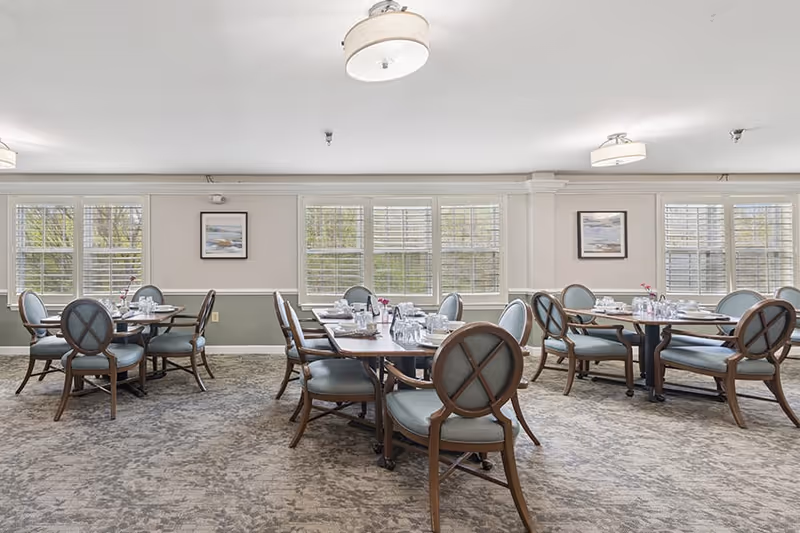 A bright dining room with multiple round wooden tables set with glasses, plates, and silverware. Each table is surrounded by cushioned chairs with wooden frames and light blue upholstery. The room has large windows with white blinds allowing natural light to fill the space. The walls are painted in a light color with a green wainscoting, and there are framed pictures hanging on the walls. The carpet is patterned in neutral tones, and ceiling lights provide additional illumination.