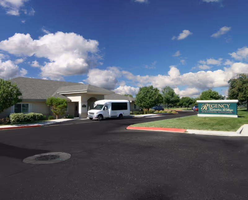 Exterior view of a senior living facility with a driveway, a white shuttle van parked near the entrance, green trees, and a sign that reads 'Regency Columbia Village Senior Living' under a partly cloudy blue sky.