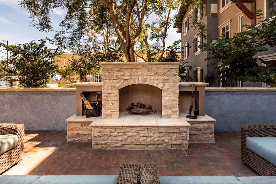 Outdoor seating area with a stone fireplace surrounded by wicker furniture with cushions, trees, and a building in the background under a partly cloudy sky.