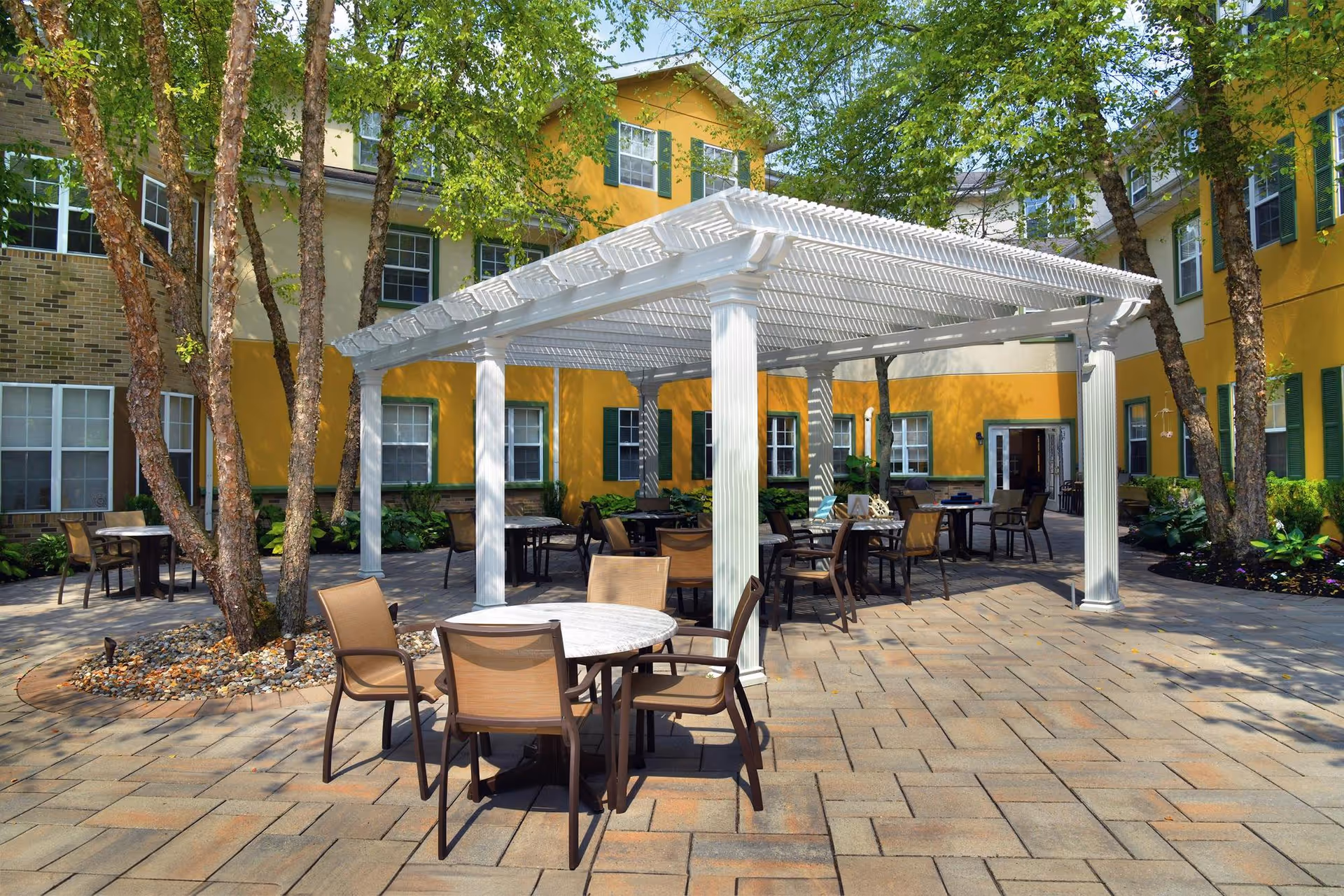 Sunlit courtyard with a white pergola, outdoor tables and chairs, trees, and a surrounding yellow assisted-living building.