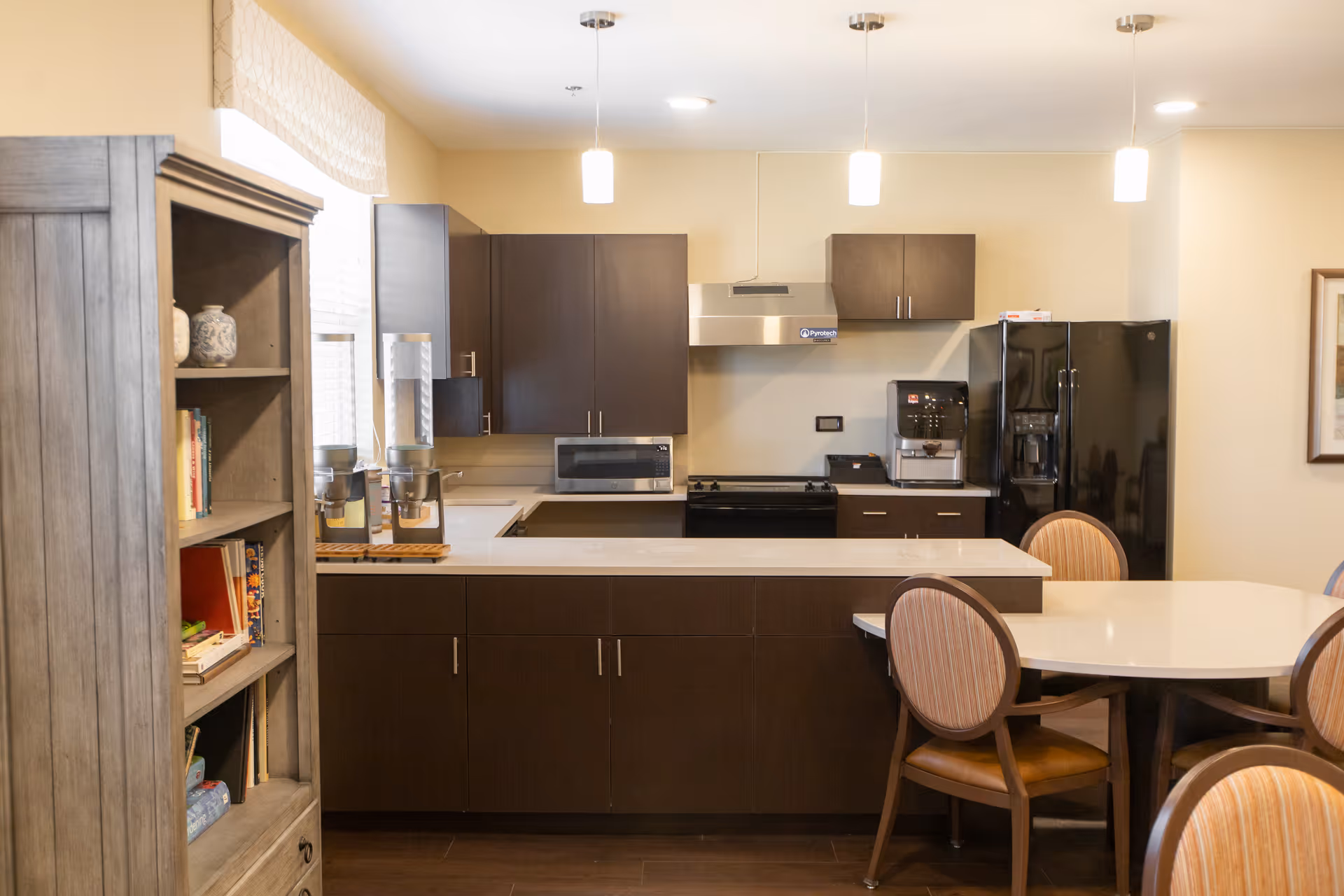 Modern communal kitchen and dining area with dark cabinets, a countertop island, appliances and a round dining table with chairs.
