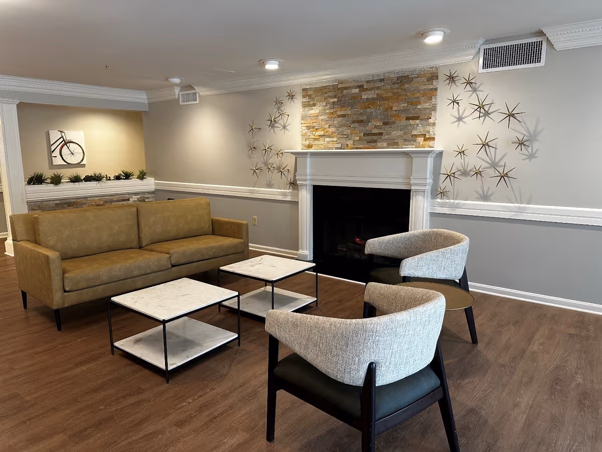 A cozy senior living facility common area with a brown sofa, two gray armchairs, two white marble coffee tables, a fireplace with a stone accent wall above it, and decorative star-shaped wall art. The room has wood flooring and neutral-colored walls with white trim.