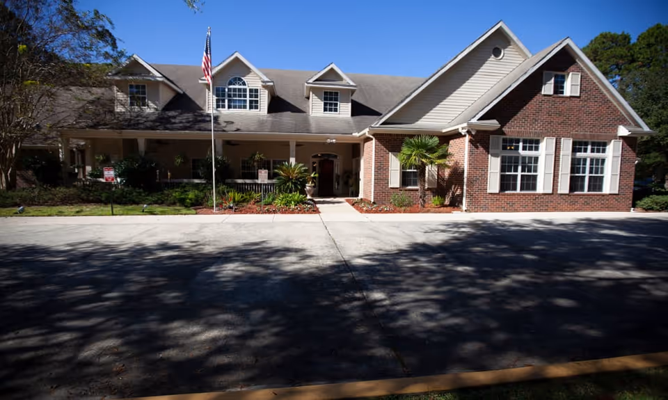 Front exterior of a brick and siding assisted living building with a porch, flagpole, and driveway under a clear blue sky.