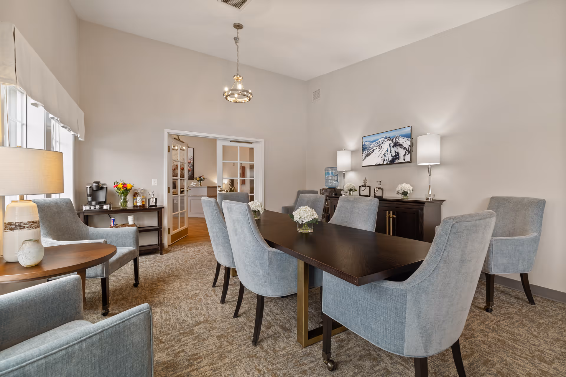 Bright dining-style room with a dark wood table surrounded by upholstered blue-gray chairs, lamps, and sideboards.