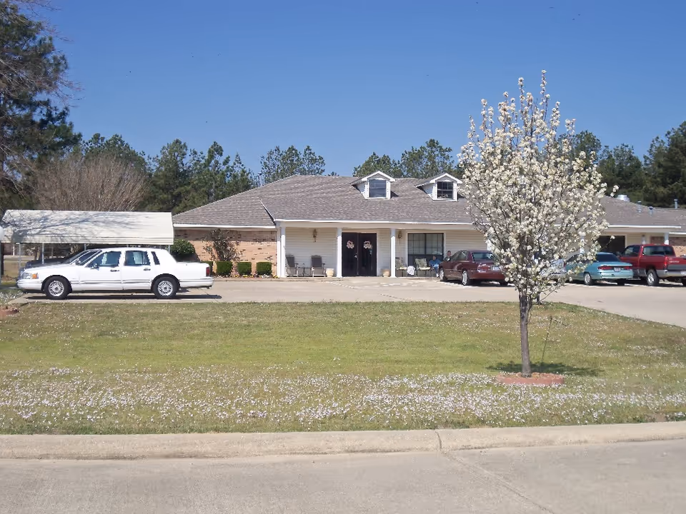 Front exterior of a single-story assisted living building with parked cars, a blooming tree, and a grassy lawn.