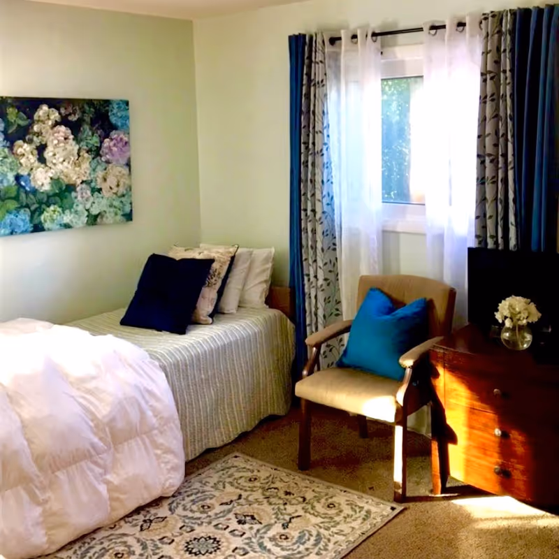 A cozy bedroom with a single bed covered in a striped bedspread and multiple pillows, including a dark blue one. Next to the bed is a beige armchair with a blue cushion. A wooden dresser with a small vase of white flowers sits beside the chair. The room has a window with sheer white curtains and patterned blue drapes. A floral painting hangs on the wall above the bed, and a patterned rug is on the carpeted floor.