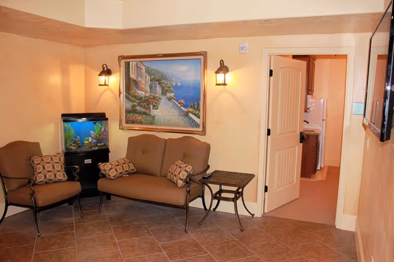 Cozy sitting area with a loveseat and chair, an aquarium, a framed seaside painting, and an open door to an adjacent room.