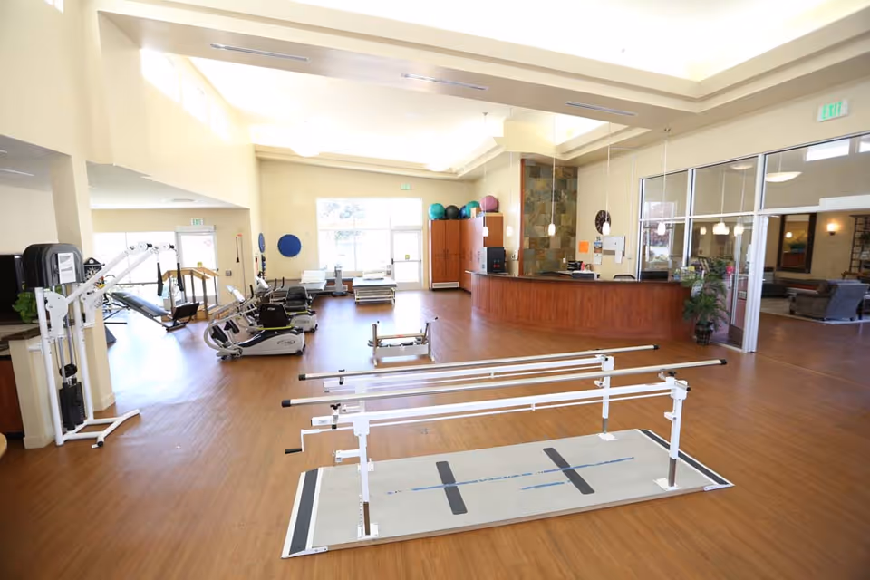 Bright rehabilitation gym with parallel bars in the foreground, exercise machines and a reception desk in the background.