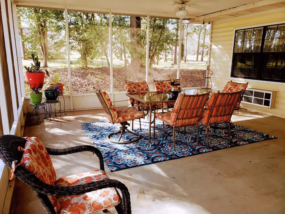 A screened-in porch area with a glass-top dining table surrounded by six cushioned chairs with floral and striped patterns. There is a patterned blue rug under the table and chairs. A wicker chair with floral cushions is in the foreground. Several potted plants are placed on metal stands near the screened walls, and trees and grass are visible outside. The porch has a ceiling fan and a window air conditioning unit on the wall.