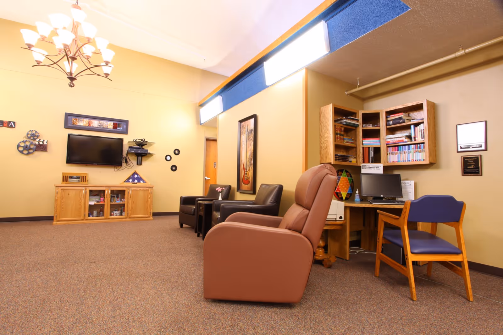 A cozy common room with armchairs, a wall-mounted television, bookshelves and a small computer desk.