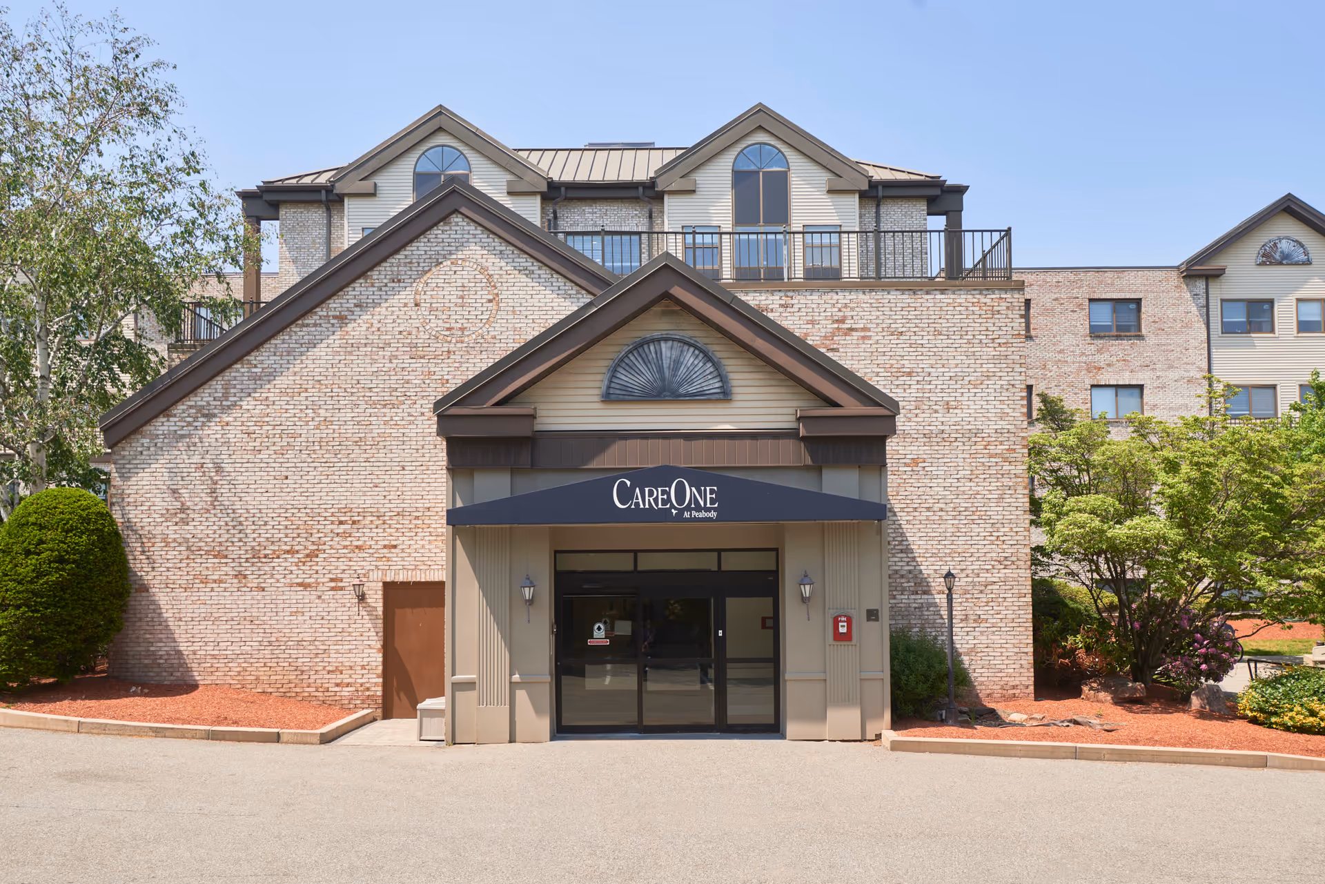 Front exterior view of CareOne at Peabody building with a brick facade, entrance canopy with the facility name, and surrounding landscaping including trees and bushes.