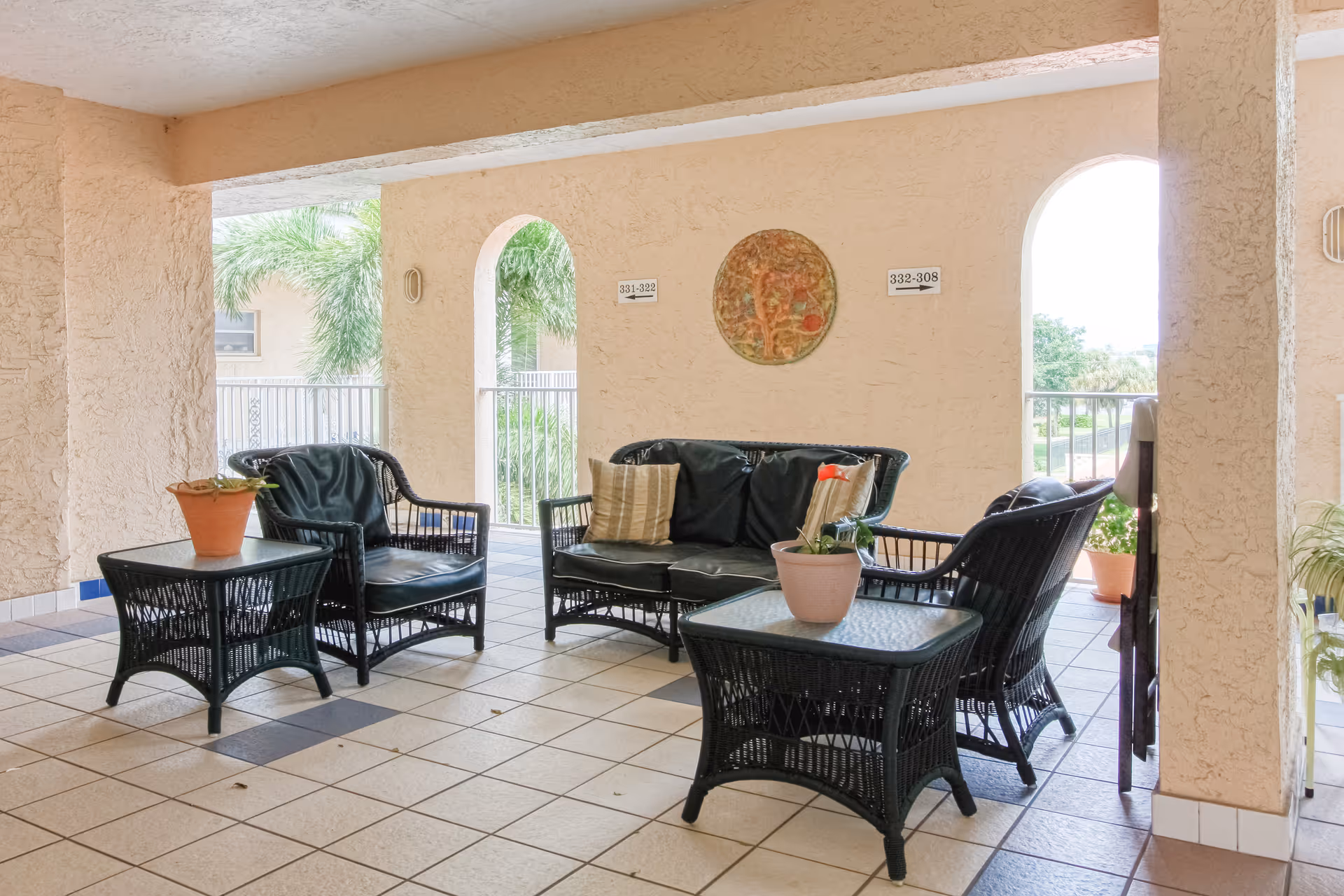 Covered outdoor seating area with black wicker furniture including a loveseat and two chairs with cushions, two glass-top wicker tables with potted plants, beige textured walls with arched openings showing palm trees outside, and tiled floor.